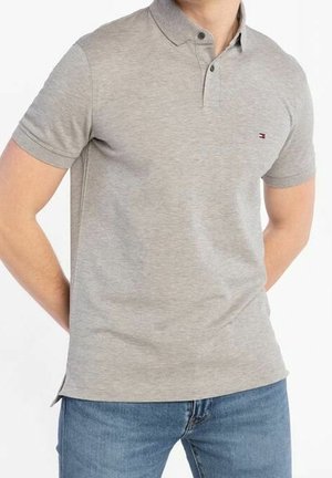 Homme portant un polo à manches courtes gris clair avec un petit logo sur la poitrine et un jean bleu, debout les mains derrière le dos.