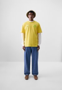 Camiseta de algodón amarilla con logo verde, superpuesta sobre una camisa de manga larga blanca. Llevada con unos vaqueros azules holgados y zapatos marrones.