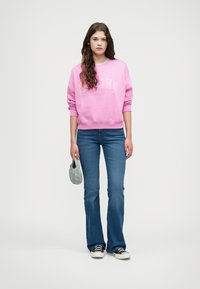 Sweat-shirt rose avec texte blanc, jeans bleus à design évasé et baskets noires. Tenant un petit sac bleu.