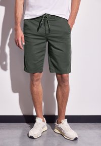 Zelené kraťasy s šňůrkovým uzávěrem, vyrobené z lehkého materiálu. Kapsy na obou stranách a volný střih. Nosené s béžovými teniskami.