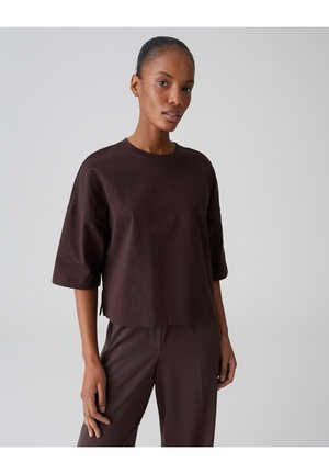 Frau mit zurückgebundenem Haar, die ein lockeres, dunkelbraunes Kurzarmoberteil und passende Hose vor einem einfarbigen hellen Hintergrund trägt.