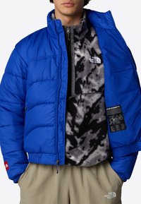 Giacca a vento blu con colletto alto e interno in felpa grigio scuro, caratterizzata da una sezione nera con motivo e toppe con logo.