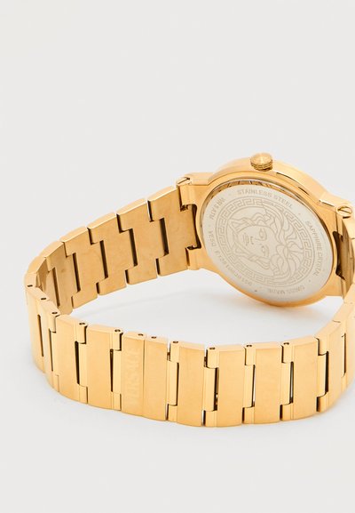 Μπαντάνα ρολογιού από μέταλλο σε χρυσή απόχρωση και πίσω θήκη με χαραγμένο κεφάλι Μέδουσας και λεπτομέρειες της μάρκας σε λευκό φόντο.