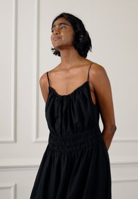 Vestido preto sem mangas com decote franzido e cintura elástica, feito de um tecido suave. O modelo tem cabelo curto e escuro e está posicionado contra um fundo neutro.