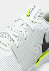 Λευκό αθλητικό παπούτσι με επάνω μέρος από αναπνεύσιμο δίχτυ, γκρι επικαλύψεις, μαύρο λογότυπο και νέον πράσινες λεπτομέρειες στη σόλα. Ορατά κορδόνια και υφή.