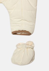 Tuta da neve soffice color crema con bordo marrone ai polsini e stivaletti abbinati color crema, caratterizzata da un design trapuntato e una texture morbida.