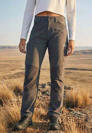 Person trägt graue Wandershosen, schwarze Stiefel und ein weißes, langärmeliges Croptop und steht auf felsigem Grasland mit offenem Landschaftshintergrund.