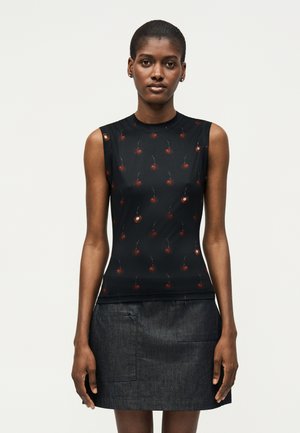 Γυναίκα με αμάνικη μαύρη μπλούζα με μικρά κόκκινα σχέδια κερασιών και σκουρόχρωμη τζιν φούστα, στέκεται μπροστά σε απλό φωτεινό φόντο.