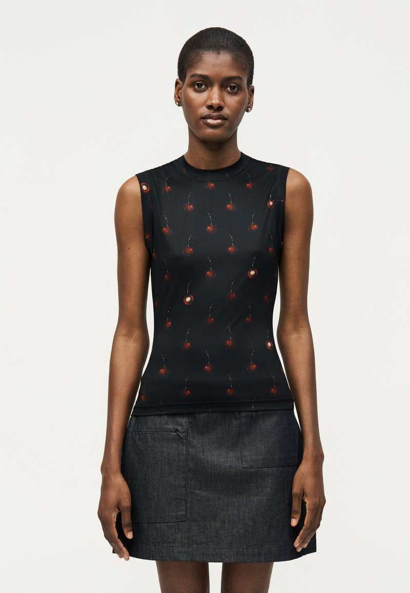 Γυναίκα με αμάνικη μαύρη μπλούζα με μικρά κόκκινα σχέδια κερασιών και σκουρόχρωμη τζιν φούστα, στέκεται μπροστά σε απλό φωτεινό φόντο.