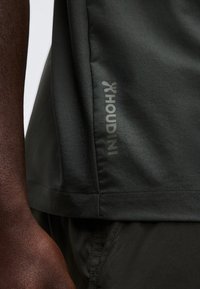 Κοντινό πλάνο ενός ατόμου που φοράει σκούρα αθλητικά ρούχα, εμφανίζοντας το πλαϊνό λογότυπο «Houdini» στο τελείωμα της μπλούζας.