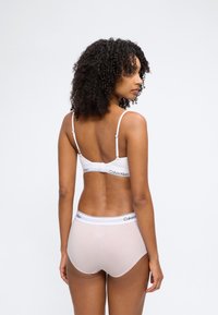 Γυναίκα με σγουρά μαλλιά που φοράει λευκό σουτιέν Calvin Klein και ανοιχτό ροζ σλιπ Calvin Klein, εμφανιζόμενη από πίσω σε απλό λευκό φόντο.