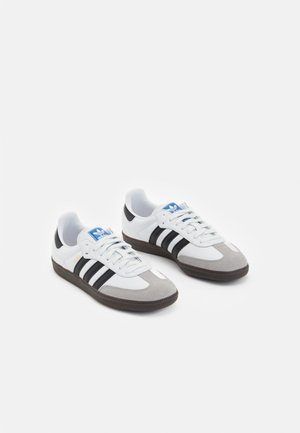 Para białych butów sportowych Adidas z czarnymi paskami, niebieską metką z logo, szarymi zamszowymi noskami i ciemnobrązowymi podeszwami na białym tle.