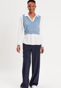Gilet en tricot bleu clair avec un col en V sur une chemise blanche à boutons, associé à un pantalon large à rayures marines et des baskets blanches.