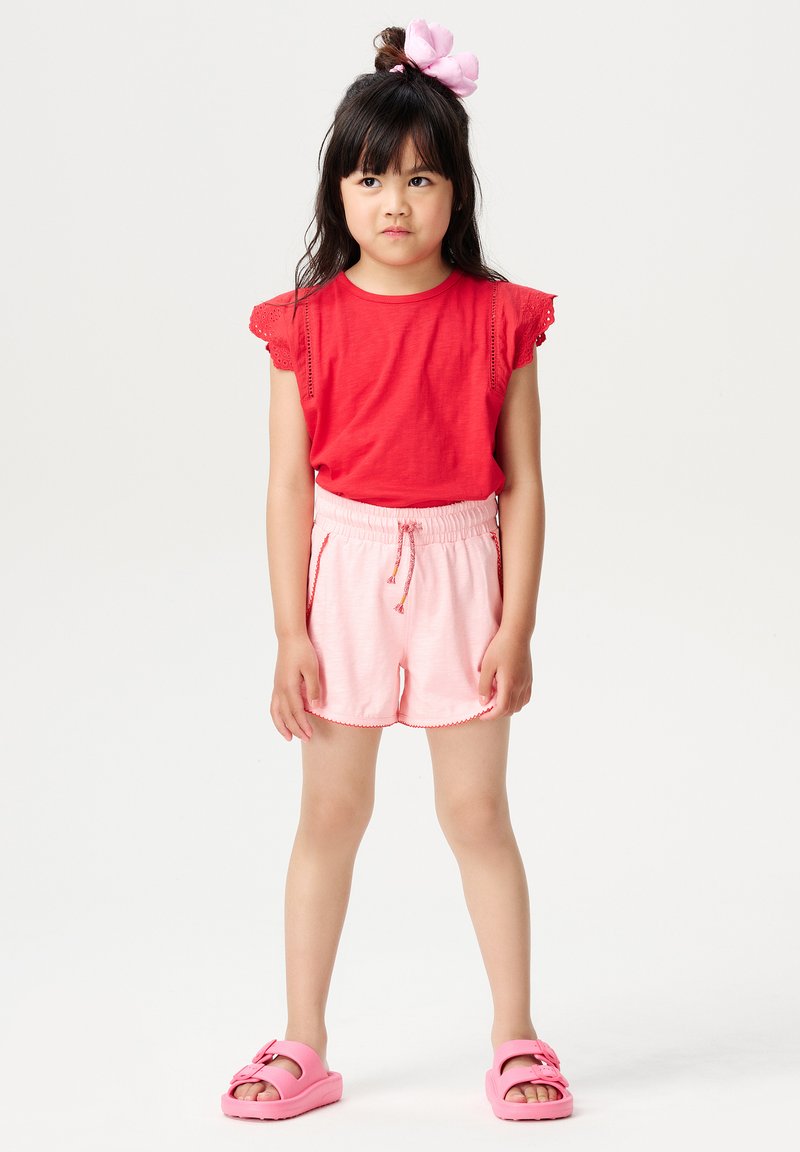 Rotes kurzärmliges Oberteil mit Spitze, kombiniert mit hellrosa Shorts mit Kordelzug und gewellten Kanten. Dazu werden rosa Sandalen getragen.
