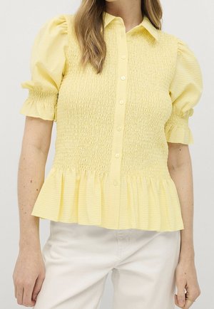 Femme portant un chemisier jaune clair à boutons, manches bouffantes, corsage smocké et ourlet à volants, associé à un pantalon blanc.