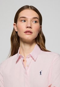 Jasnoróżowa koszula zapinana na guziki z klasycznym kołnierzykiem i małym granatowym logo przedstawiającym golfistę po lewej stronie na piersi. Gładka struktura materiału.