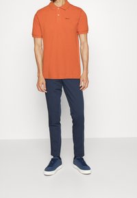 Oranžové bavlněné polo tričko s krátkými rukávy, límečkem a černým logem. V kombinaci s námořnickými kalhotami a modrými teniskami s bílou podrážkou.