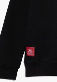 Czarna bluza z prążkowanymi mankietami, z czerwoną metką na rękawie z logo i napisem. Materiał wydaje się miękki i elastyczny.