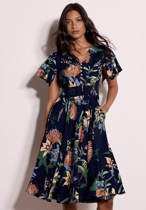 Frau mit langen dunklen Haaren, die ein marineblaues, geblümtes Midikleid mit kurzen Rüschenärmeln und Knöpfen am Oberteil trägt.