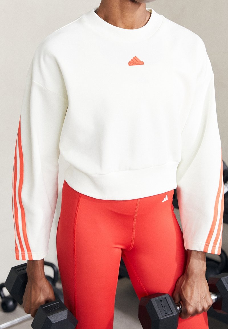 Bílá mikina s oranžovým logem Adidas a třemi oranžovými pruhy na rukávech, spárovaná s těsnými červenými legínami, držící pár činek.