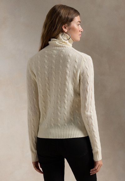 Femme portant un pull à col roulé en maille torsadée ivoire et un pantalon noir, de profil, avec de longs cheveux bruns et de grandes boucles d'oreilles.