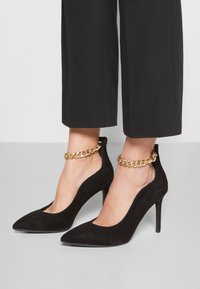 Svarta mocka högklackade skor med spetsiga tår och tunna stilettklackar. Har guldkedjedetalj runt vristen.