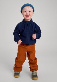 Giacca in pile blu navy, pantaloni morbidi arancioni e berretto di maglia blu. Sneakers grigie con suola beige. Bambino in posa con un sorriso.