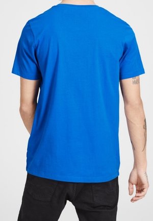 Osoba ubrana w gładki, jasnoniebieski t-shirt z krótkim rękawem i czarne spodnie, widziana od tyłu, z widocznymi tatuażami na ramionach.