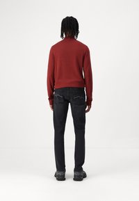 Maglione rosso a maglia con colletto alto, abbinato a jeans grigio scuro con dettagli in bianco sulle tasche posteriori. Sneakers nere.