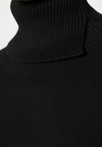 Pull à col roulé en maille côtelée noire en gros plan montrant la texture et le pli au niveau de l'encolure.