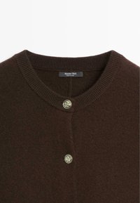 Brązowy dziergany kardigan z okrągłym dekoltem, z zapięciem na guziki i okrąłym metalowym akcentem. Miękka tekstura i minimalistyczny design.