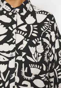 Svart och vit skjorta med knappslå, prydd med ett blommigt och abstrakt mönster, med en bröstficka och svarta knappar. Mjuk tygkänsla.