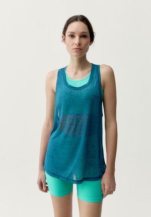 Ung kvinde iført teal sports-bh, løs blå ærmeløs top og teal cykelshorts, stående mod en ensartet lys baggrund.