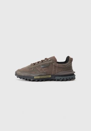 Baskets basses Lacoste marron avec semelle noire texturée, lacets à l'avant et panneau latéral cousu sur fond gris clair uni.