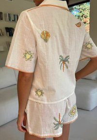 Baltas lininis marškinėlių ir šortų komplektas su spalvinga gėlių ir kriauklių siuvinėjimu, oranžiniu apvadu ir laisvu kirpimu.