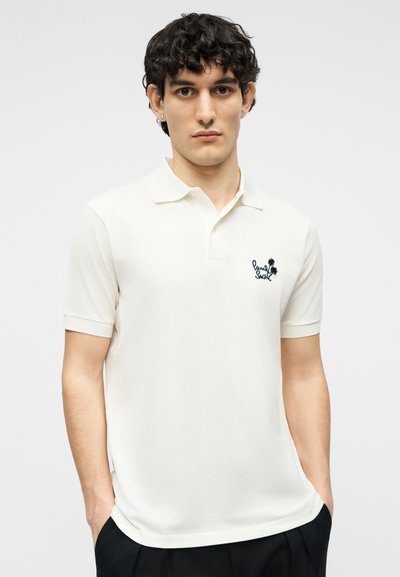 Νέος άνδρας με σγουρά σκούρα μαλλιά που φοράει λευκό λινό πόλο με κοντά μανίκια και μικρό μαύρο κεντημένο λογότυπο στο στήθος.