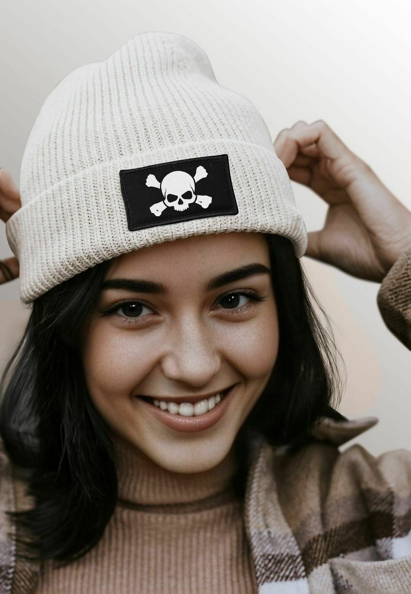 Beanie hat in off-white ribbed knit, featuring a black patch with a white skull and crossbones design. Soft texture and fitted shape.