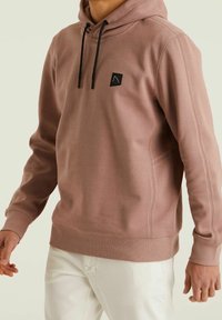 Felpa marrone in morbido tessuto con cappuccio regolabile, polsini a coste e un piccolo emblema nero sul petto. Abbinata a pantaloni bianchi.