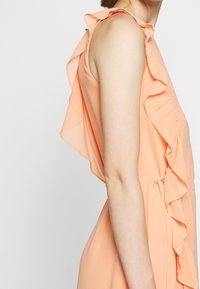 Robe couleur pêche avec détail à volants sur les épaules, en tissu léger et taille cintrée. Présente une texture lisse et un design fluide.