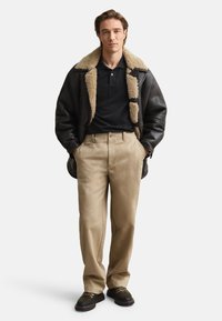 Hombre con chaqueta de cuero con forro de lana negra, camiseta polo negra, pantalones beige y zapatos negros, de pie con las manos en los bolsillos.