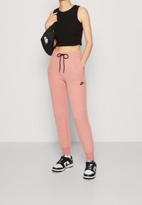 Světlé růžové tepláky s černou šňůrkou a logem Nike, spárované s černým crop topem a černobílými teniskami.