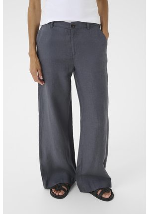 Personne portant un pantalon ample gris foncé, un haut blanc et des sandales tressées noires, debout avec les mains dans les poches sur un fond clair.