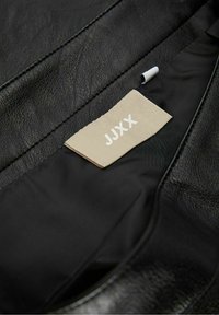 Material de cuero negro con una superficie texturizada. Cuenta con una etiqueta beige que muestra "JJXX" en blanco, situada contra un forro interior negro.