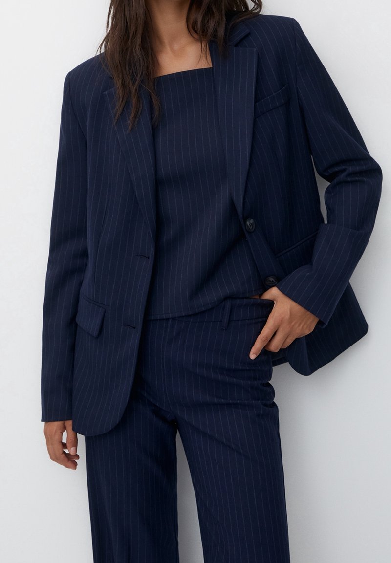 Femme portant un costume bleu marine à fines rayures avec un blazer, un haut assorti et un pantalon, debout devant un fond blanc uni.