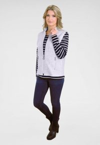 Grau strukturiertes Weste über einem schwarz-weiß gestreiften Langarmshirt, kombiniert mit dunklen Jeans und schwarzen Stiefeletten. Reißverschluss vorne mit Zuglaschen.