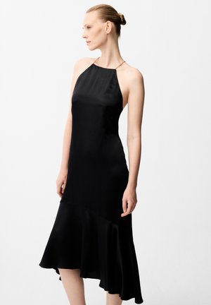 Vrouw met haar in een knot, gekleed in een mouwloze zwarte halter midi-jurk met een uitlopende asymmetrische zoom, kijkend naar links tegen een effen achtergrond.