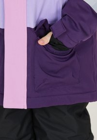 Lila und lavendelfarbene Jacke mit einem pinken Streifen, ausgestattet mit einer Tasche auf der linken Seite, aus glattem Stoff mit sichtbaren Nahtdetails.