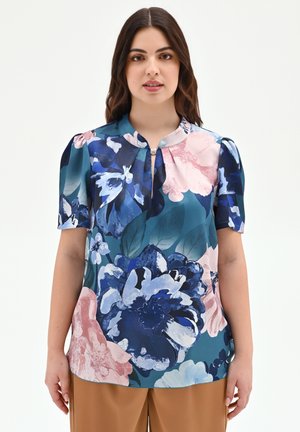 Donna che indossa una blusa turchese con grandi motivi floreali rosa, blu e bianchi e pantaloni beige, in piedi davanti a uno sfondo bianco semplice.