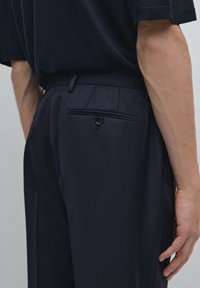 Navy pinstriped trousers featuring a slim fit, smooth texture, and a single back pocket with button closure. Minimalist design details evident.
