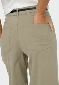 Personne portant un pantalon beige avec une ceinture noire, la main droite placée dans la poche arrière, sur un fond blanc.